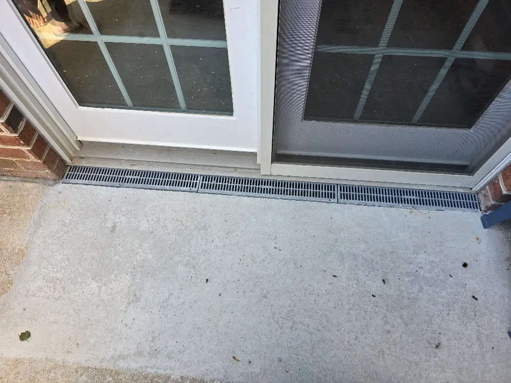 A glass double door with grid panels opens onto a concrete patio. A narrow, metal drain is positioned at the base, ensuring water drainage.